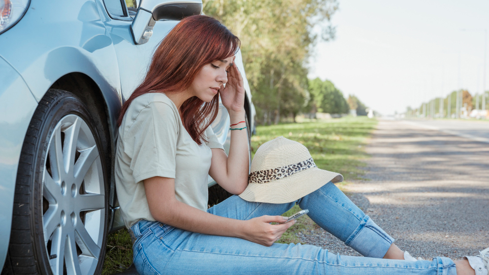 young redhead woman sitting on the side of the road on her phone after she crashed friend's car