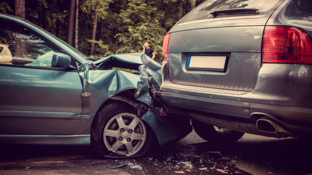 Rear-end collision in a parking lot accident, showing a teal sedan with a crumpled front end pressed into the back of a gray SUV with debris scattered on the pavement.